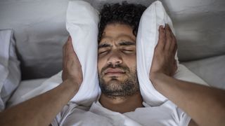 A man with dark curly hair holds a bed pillow against his ears to block noise that has woken him up from sleep