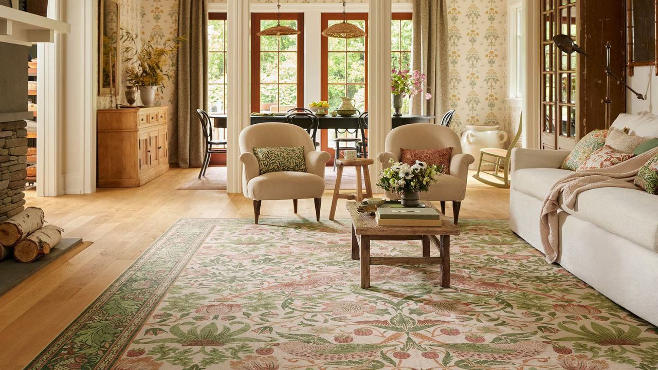 A pink, green and cream floral rug on the floor, surrounded by a white sofa and two cream armchairs; in the background, floral wallpaper and windows with a garden view.