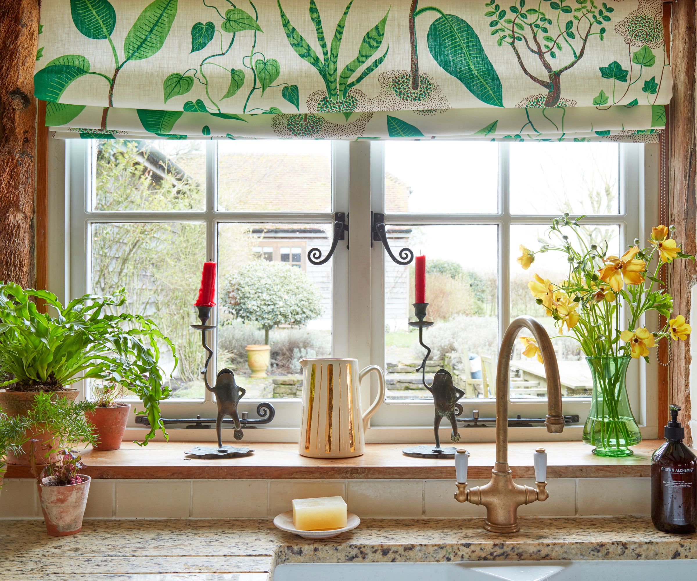A cottage kitchen window with green and white botanical print fabric for the blinds, and glass and ceramic decor on the window sill