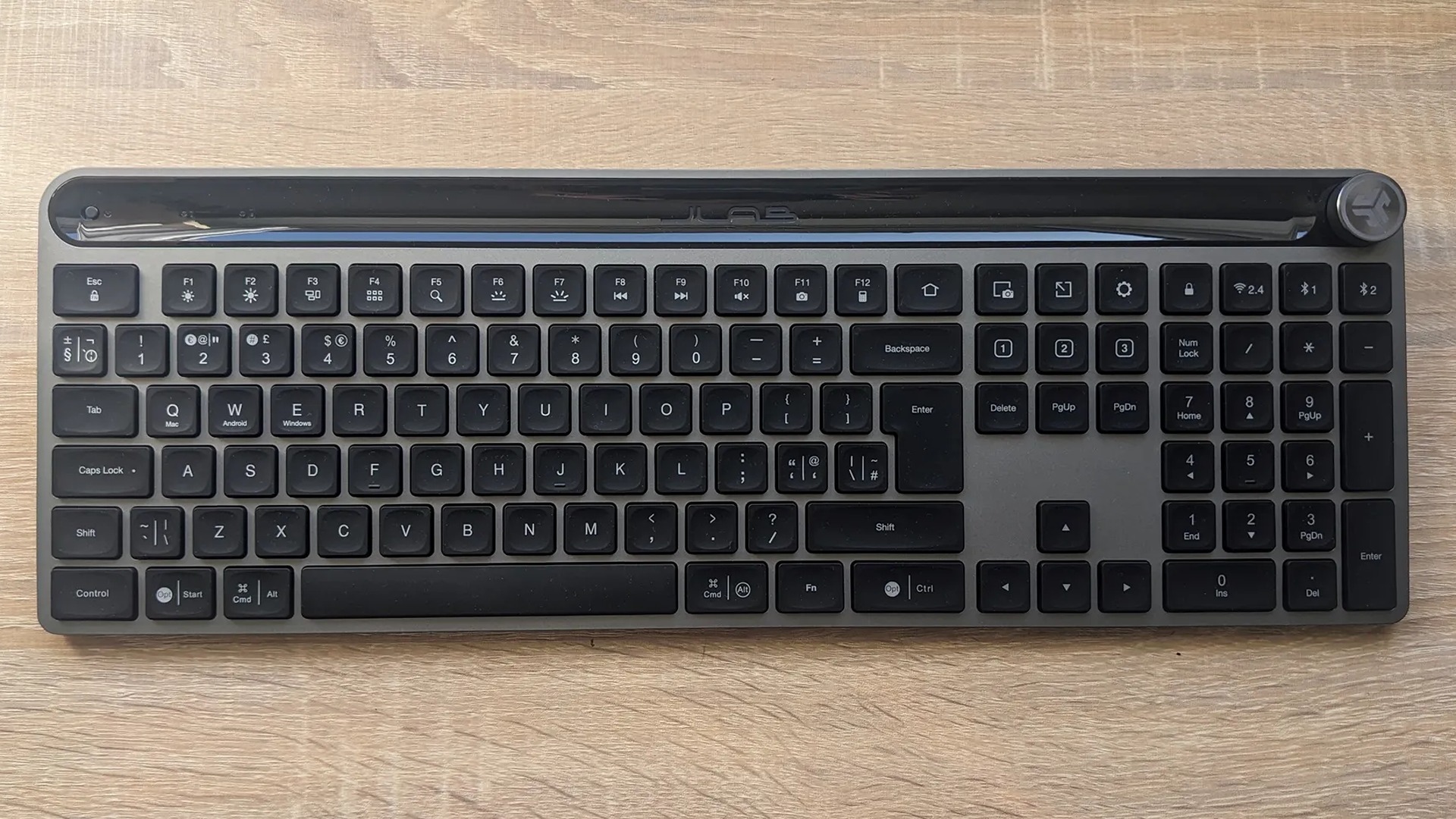 A dark gray wireless computer keyboard with a full numeric keypad and a volume control dial on a light wood grain desk surface, viewed from above.