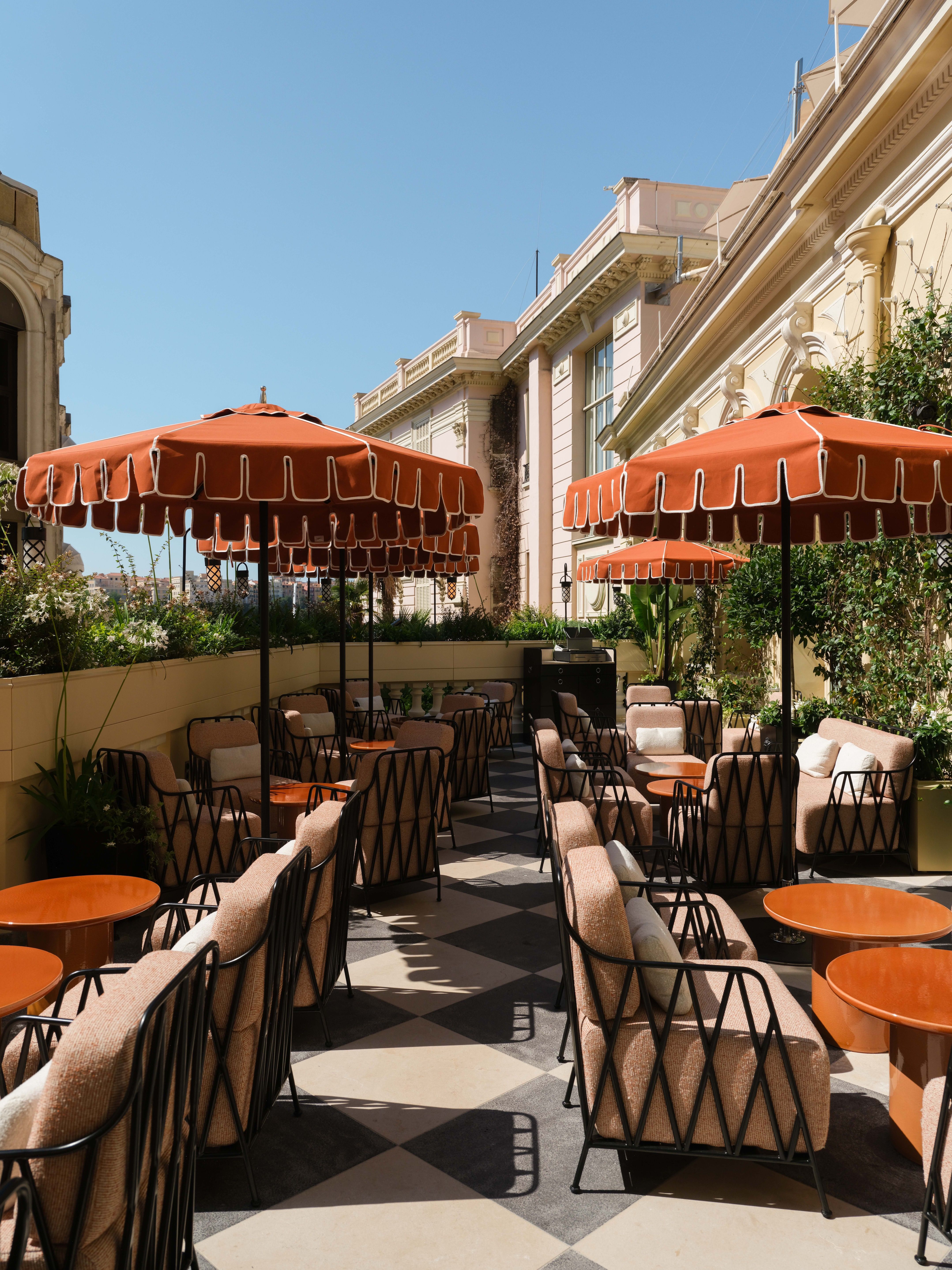 An outdoor terrace in Monaco with black and white pavers, plump outdoor seats, and burnt-orange scalloped parasols. 