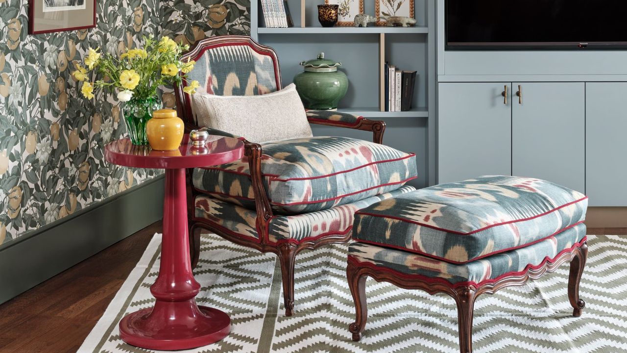 Intriguing patterned living room with a large, tall, blue media unit, classic armchair and foot-rest with brown wood and blue, cream and red upholstery, as well as a red side table. There is a geometric patterned gray and white rug, hardwood floors and patterned floral green wallpaper
