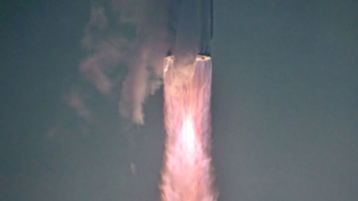 a giant finned rocket rises from glowing smoke next to a dark launch tower in a hazy setting.