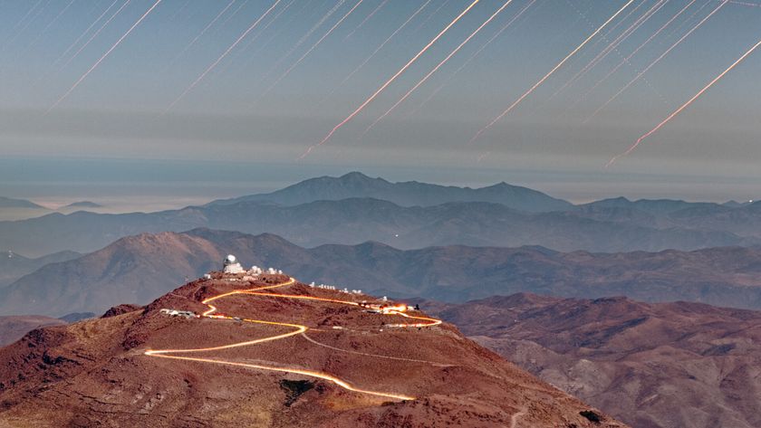 A large mountain is seen with a glowing windy road leading up it to two buildings at the top, with the rest of the rocky desert landscape all around and red streaks in the sky above.