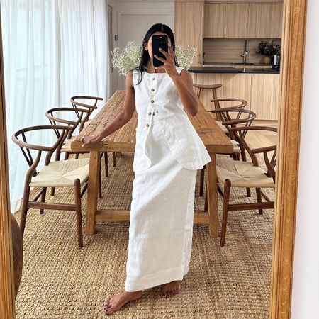 British fashion influencer Monikh Dale poses for a mirror selfie in front of her chic rustic wooden dining table, wishbone dining chairs, and jute rug wearing a white linen set with a long vest and maxi skirt