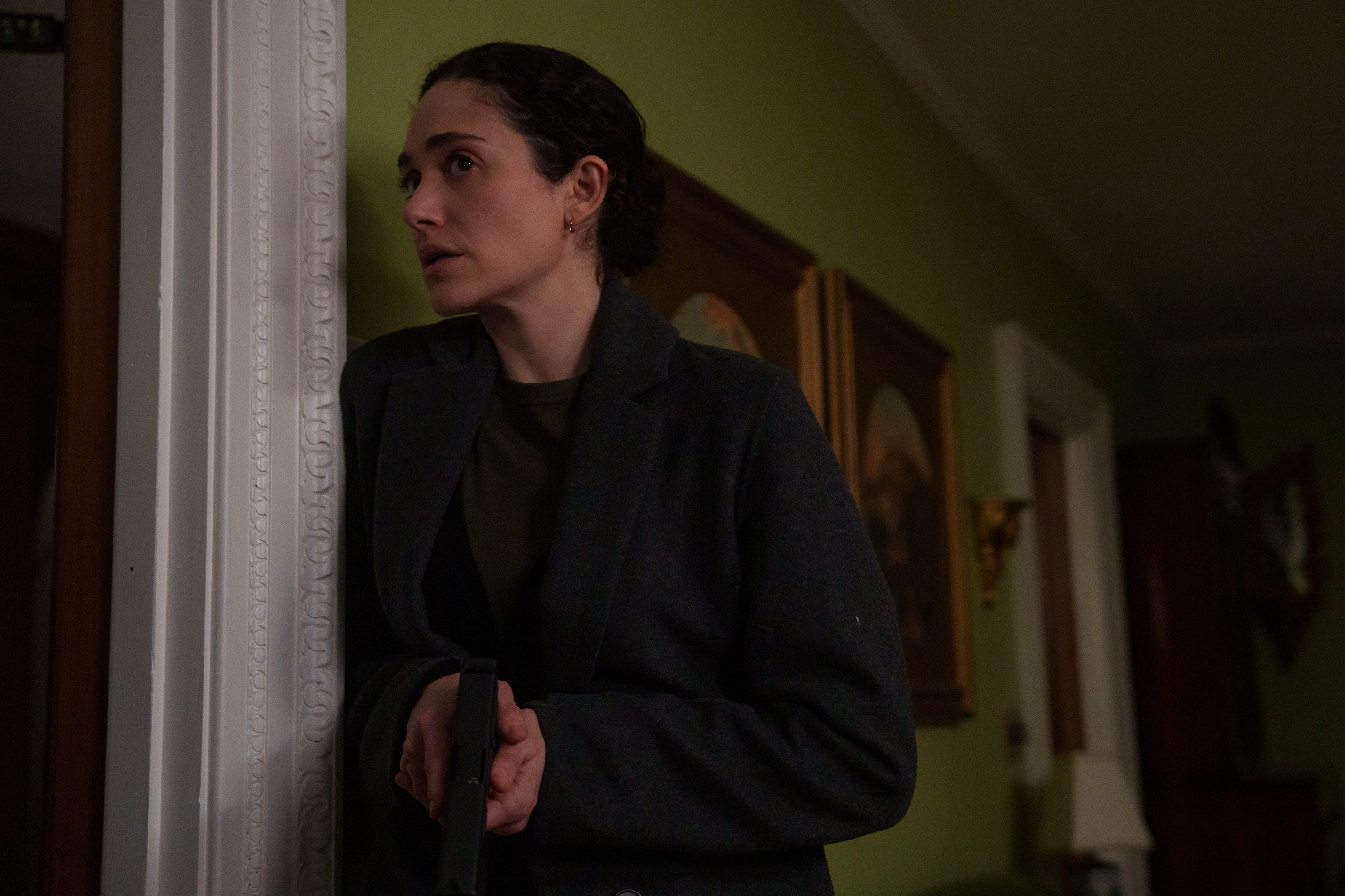 A woman (Emmy Rossum as Alice) leans against a home's hallway while holding a gun, in Hulu's 'Furious.'