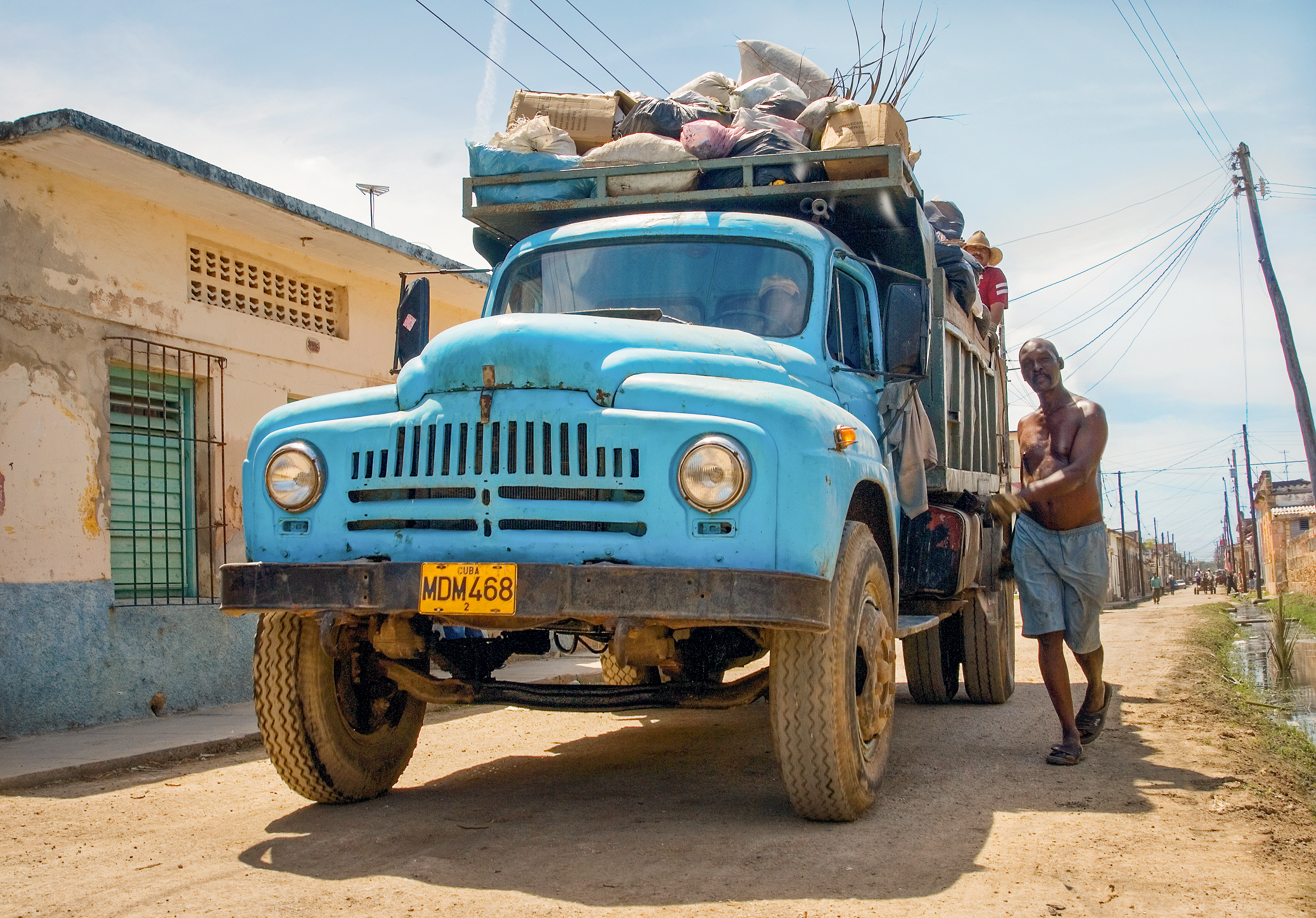 A man standing next to a heavily-loaded blue pickup truck, in a Cuban street