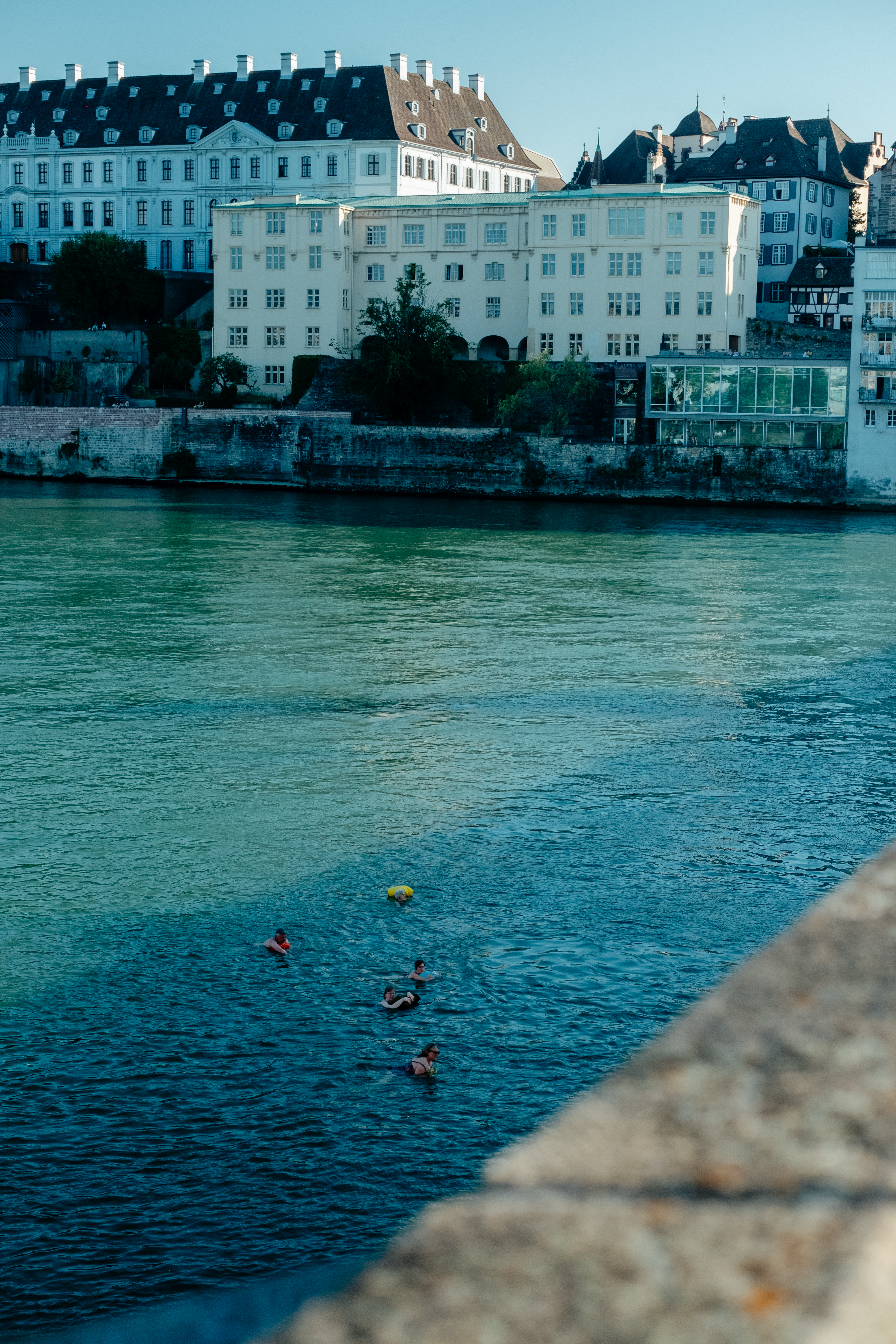 A series of lifestyle images depict people at leisure and play in a Swiss town bathing in sunshine, whether enjoying the weather near the river bank or abstract painting murals at an art fair.