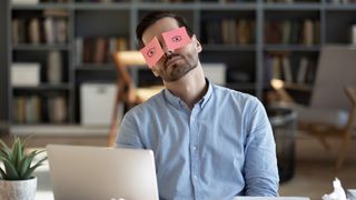 A man asleep at his desk with sticky notes over his eyes that have eyes drawn on them