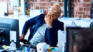 The image shows a man rubbing his eyes while sat at his desk