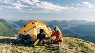 Two people enjoying a hot drink outside a tent on top of a mountain.