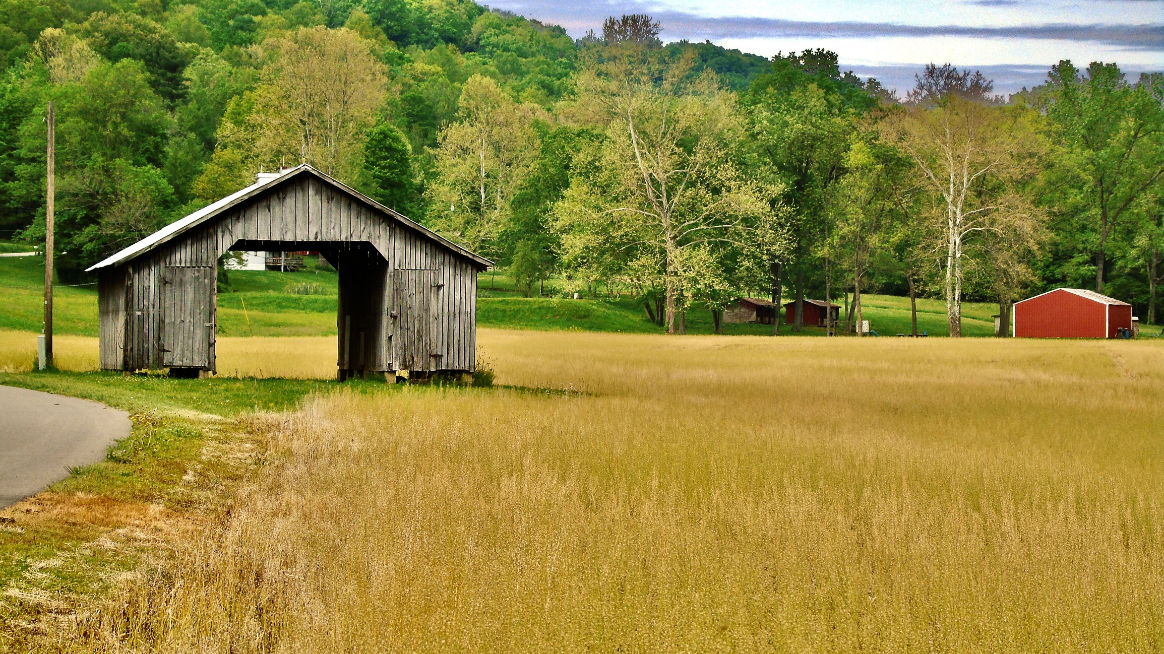 a wooden barn is beside a street, on the edge of a golden field with rolling hills and a red barn in the background, photographed in Pike County, Ohio