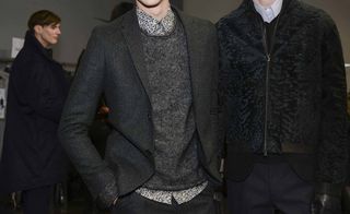 Three male models wearing looks from Fendi's collection. The close up view shows two models wearing grey, blue and black trousers, sweaters and jackets and light coloured shirts. And the third model is in the background wearing a long dark blue coat