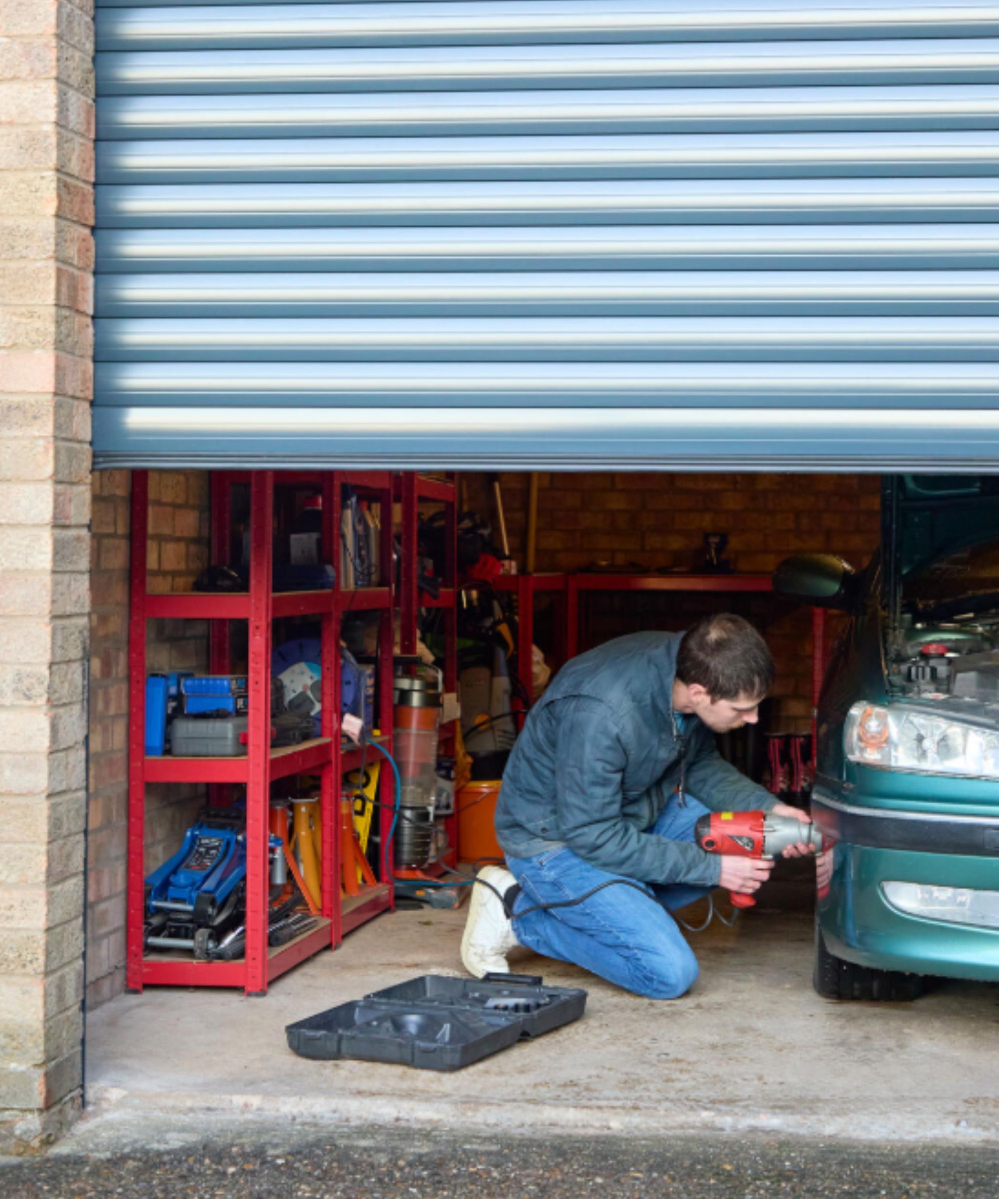 A man kneels on the concrete floor of a brick-walled garage with a partially open blue roller door. He’s using a power tool to work on the front of a green car. Red metal shelving in the background is stocked with tools and equipment, creating a tidy DIY workspace.