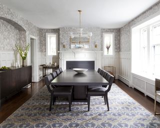 A dining room with white panelled lower walls, neutral wallpaper, black dining table and brass chandelier
