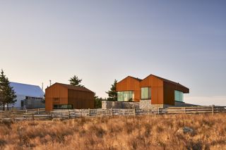 Alternative exterior view of Smith House during the day which is made up of three Corten steel and stone pavilion structures. The pavilions feature large windows and pitched roofs. There are trees and brown grassy land nearby