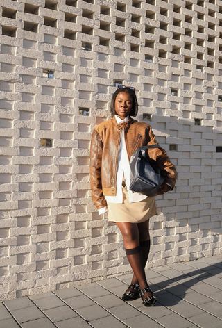 A woman wearing a tan moto jacket with a white button-down shirt, a tan skirt, sheer socks, and ballet flats.