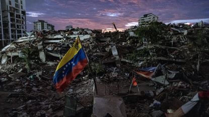 A view of the aftermath of an earthquake, with rubble everywhere and a red, yellow and blue flag in the center.