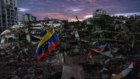 A view of the aftermath of an earthquake, with rubble everywhere and a red, yellow and blue flag in the center.