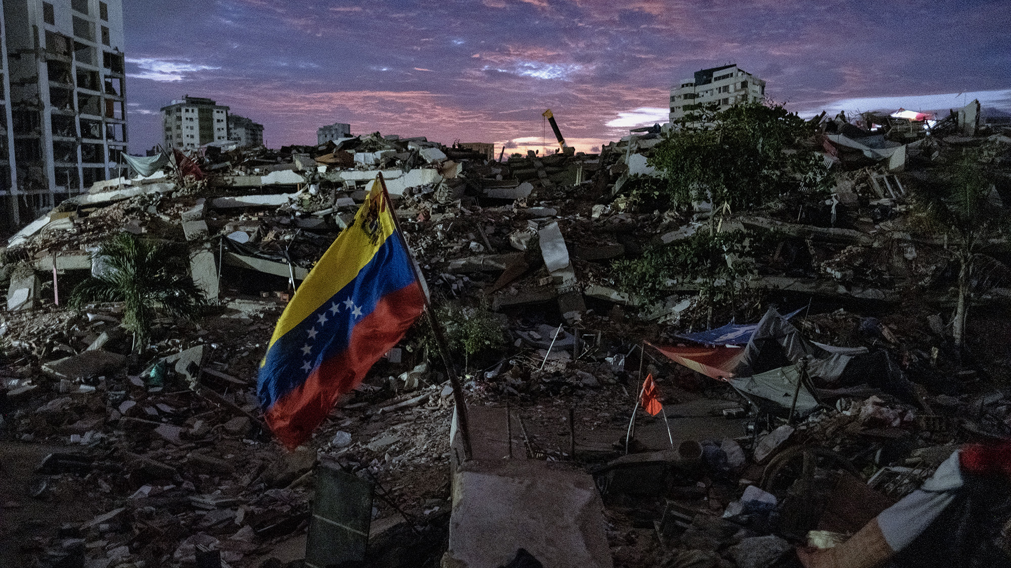 A view of the aftermath of an earthquake, with rubble everywhere and a red, yellow and blue flag in the center.