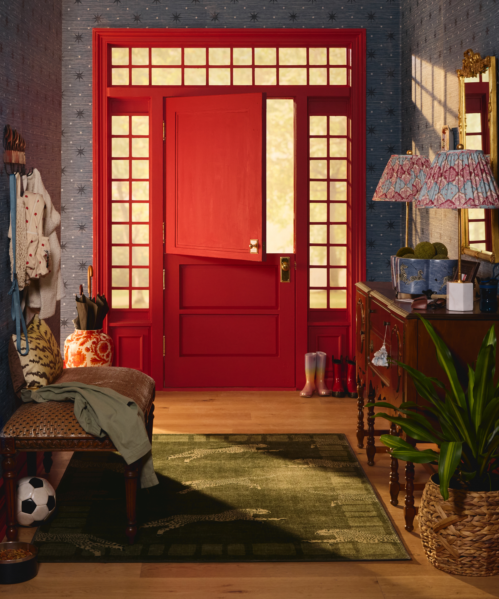 an entryway with a bright red dutch door, blue wallpaper, a busy entryway console table with two lamps on top, and an entryway rug