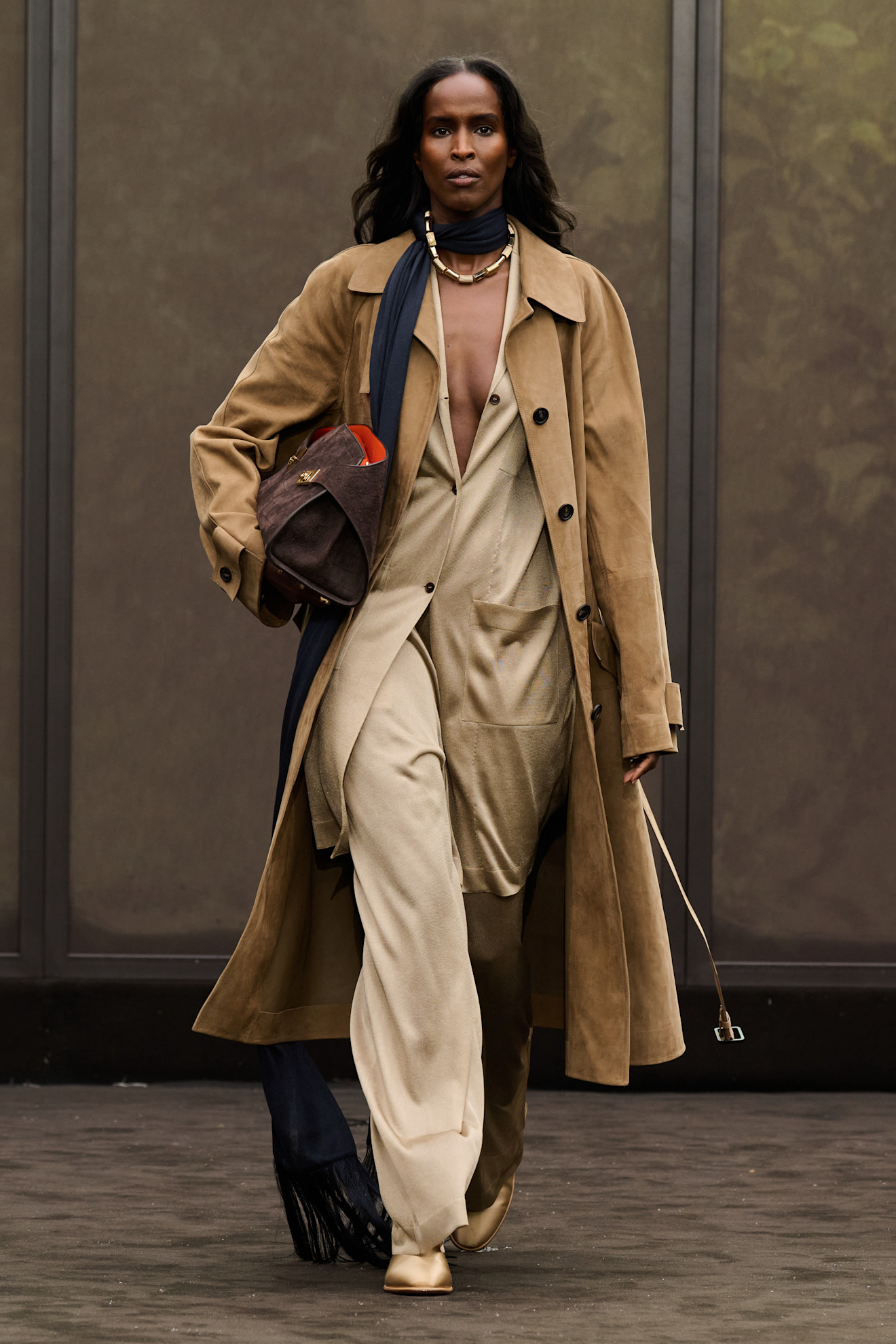 Layering staples for spring 2026 are shown in a photo from Ferragamo's runway show, with a model walking while wearing a tan suede trench coat layered over a tan duster cardigan with tan wide-leg trousers, tan satin pointed pumps, a long black fringe scarf, a gold necklace, and a brown suede clutch bag