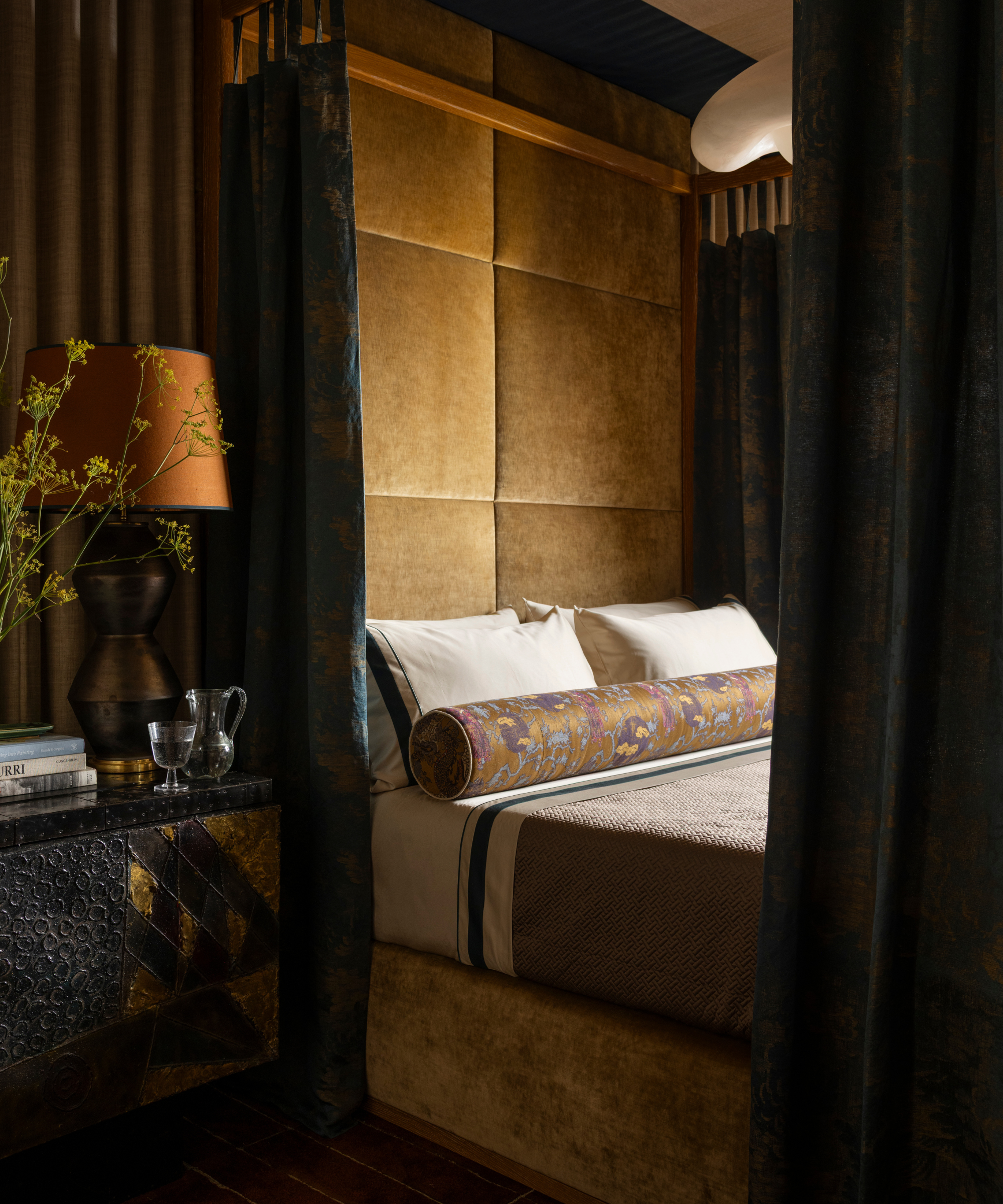 Opulent bedroom vignette featuring a canopy bed with an oversized velvet headboard, flanked by jacquard drapes, styled with piped bordered bed sheets, textured coverlet, and patterned lumbar pillow.