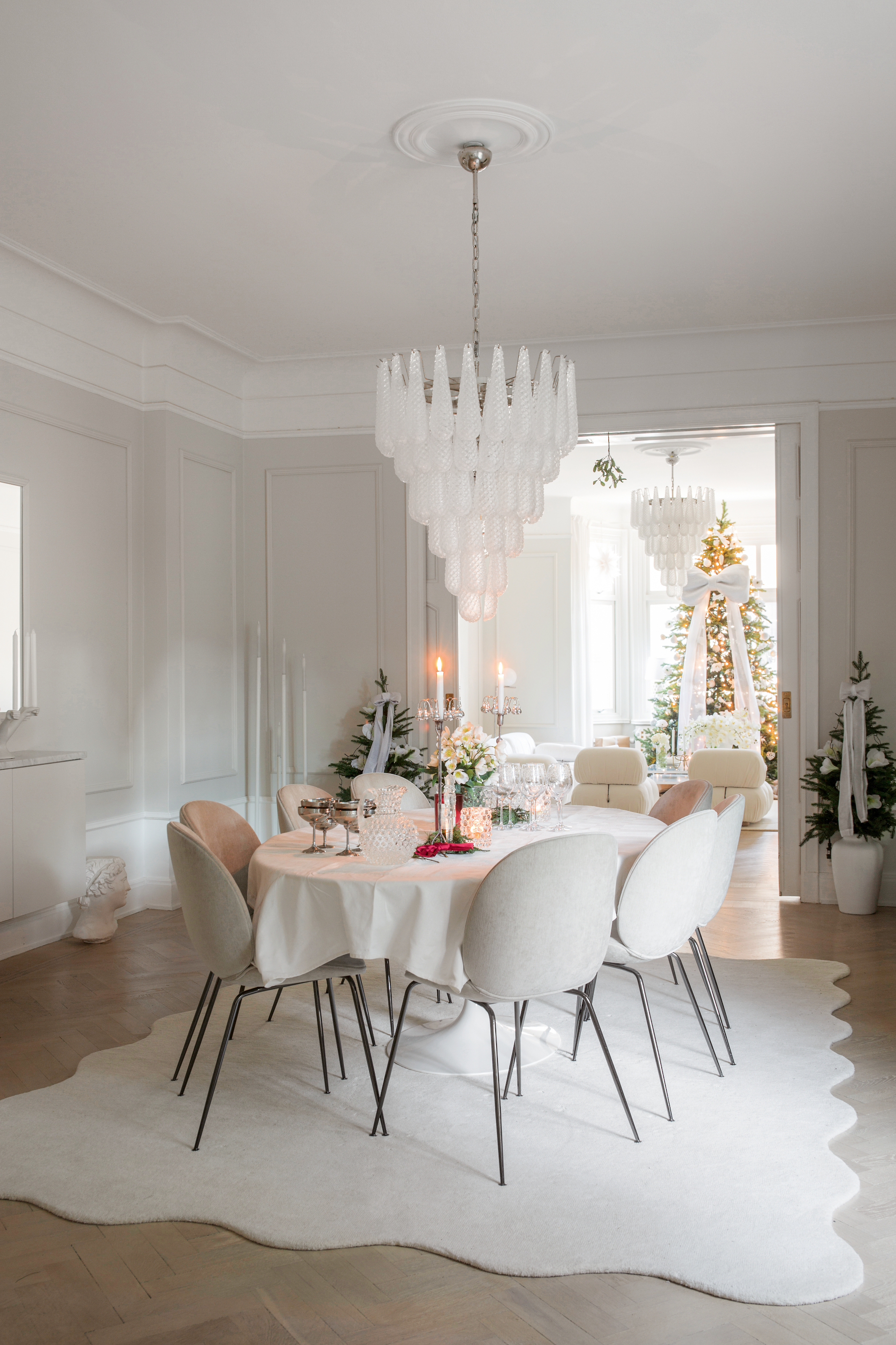 White dining room with white-covered table, white chairs, white rug and white glass chandelier