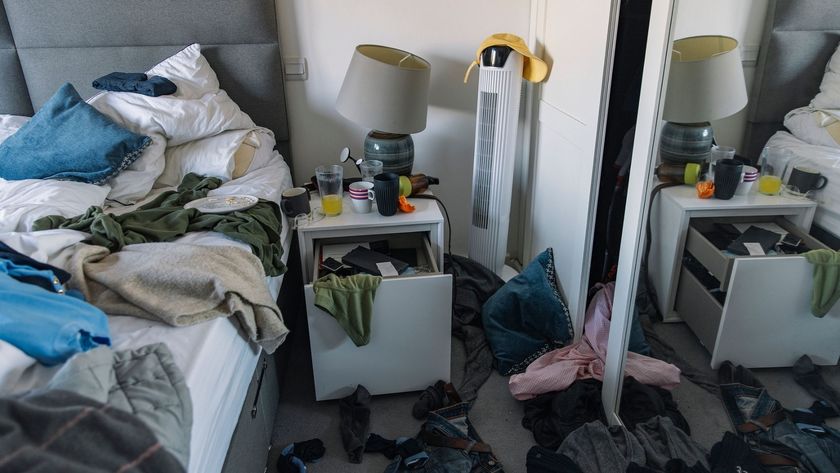 The image shows a messy and cluttered bedroom, with the bed unmade, clothes on the bed and floor and the bedside table drawers left open