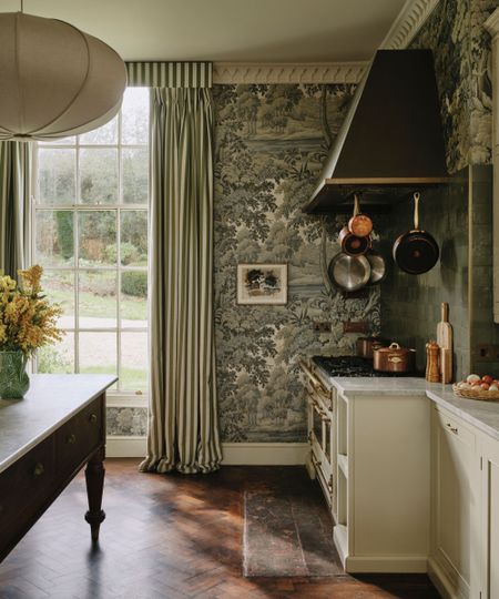 A kitchen in a traditional home with a nature-inspired wall mural in sage green tones. A floor-to-ceiling window with green striped curtains, dark wood parquet flooring, off-white cabinets, and copper pans above the oven.