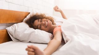 A smiling woman with brown curly hair lies in a bed covered with a white comforter
