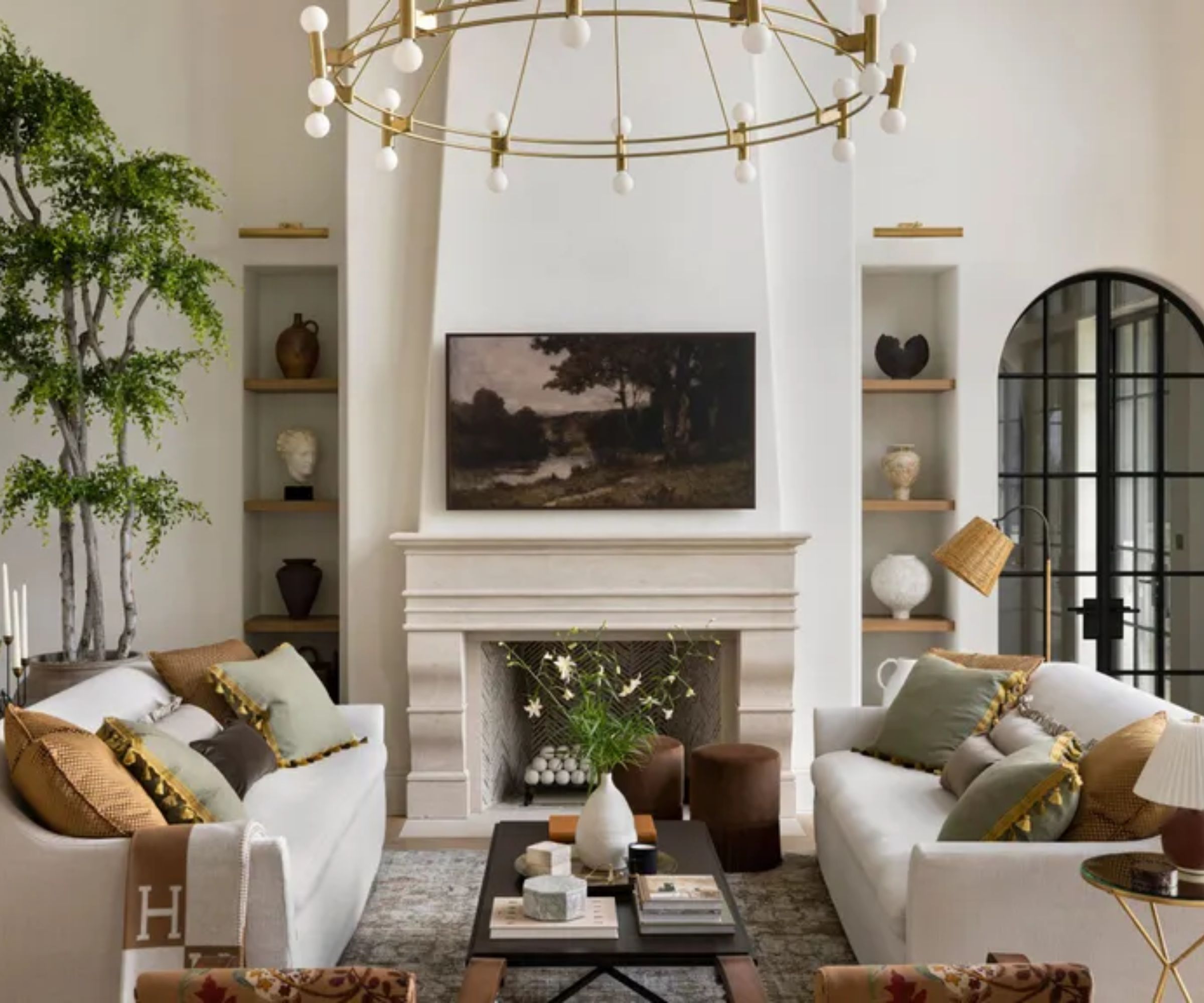 A bright living room with two white sofas facing a stone fireplace. A large tree sits in the corner, and shelves with pottery line the fireplace. A round gold chandelier hangs above, and tall black-framed windows let in natural light. A coffee table with books and flowers sits in the center.