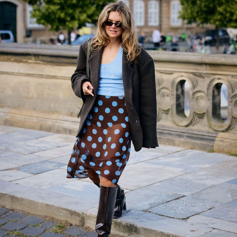 A woman wears a light blue tank, blue polka dot skirt, blazer, and tall boots. 