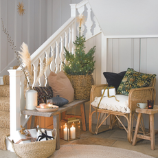 Hallway and staircase with a basket of wrapped presents, rattan rug, chairs and small decorated Christmas Tree with small table with lit candles.