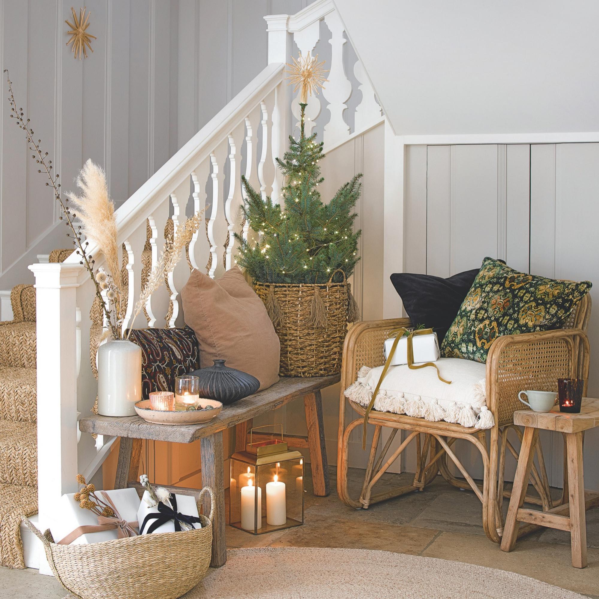 Hallway and staircase with a basket of wrapped presents, rattan rug, chairs and small decorated Christmas Tree with small table with lit candles.