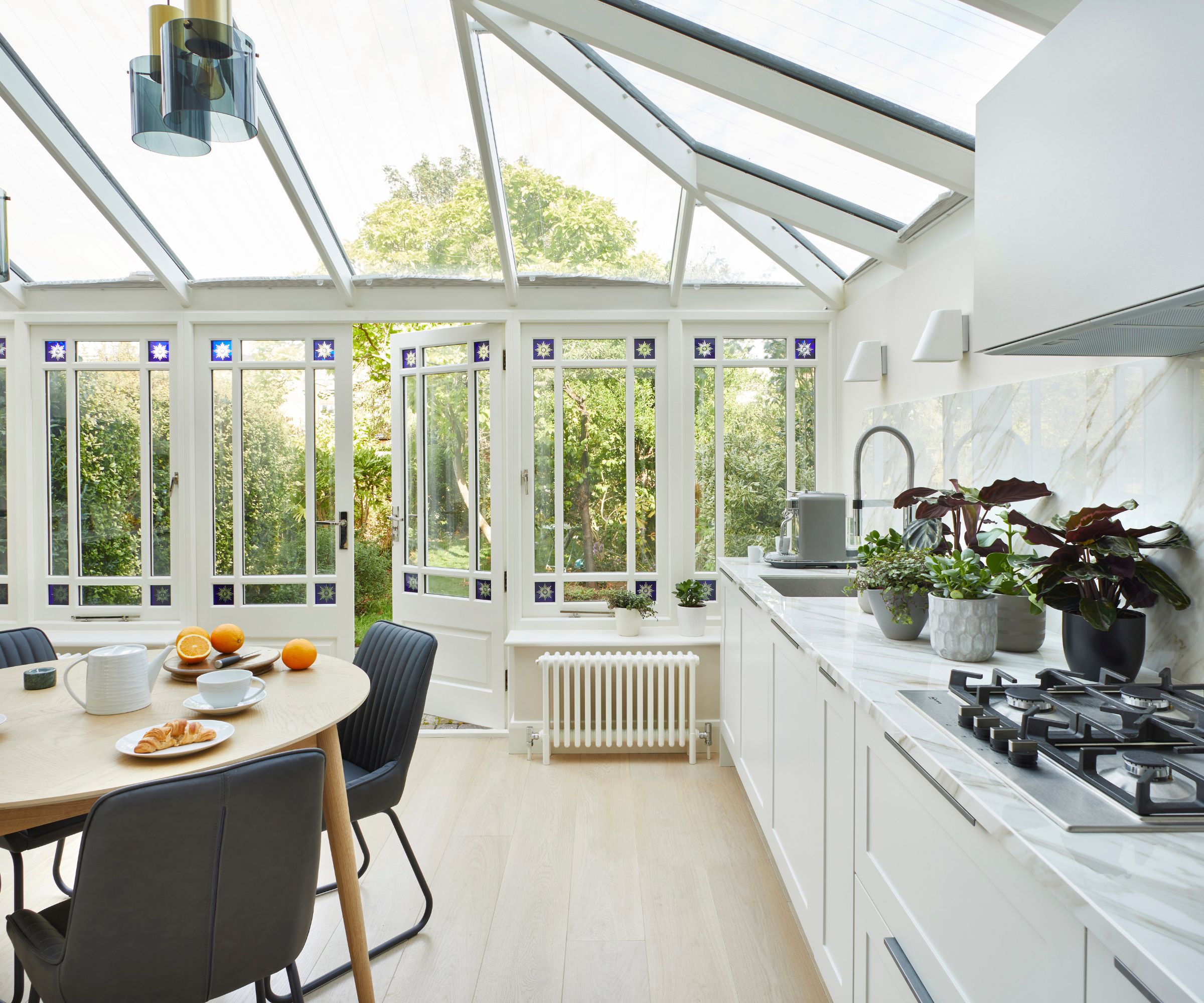 A bright white kitchen-diner in a Victorian conservatory with a pitched glass roof, blue stained glass window details, marble worktops, a gas hob, and a round oak breakfast table.