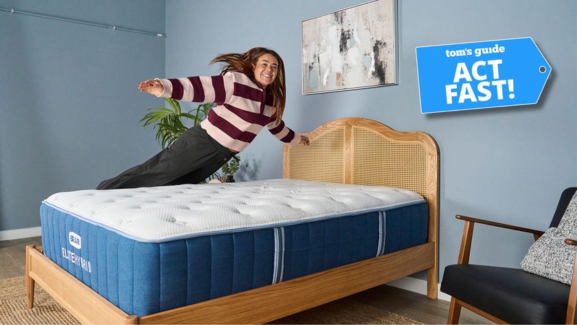 Our mattress tester jumping on the Bear Elite Hybrid mattress on a wooden bedframe with a rattan headboard in our sleep studio with blue walls and a bedside plant with Tom's Guide 'Act Fast' logo