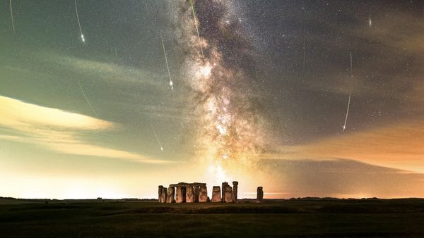 The silhouette of stonehenge is seen under a starry sky with shooting stars.