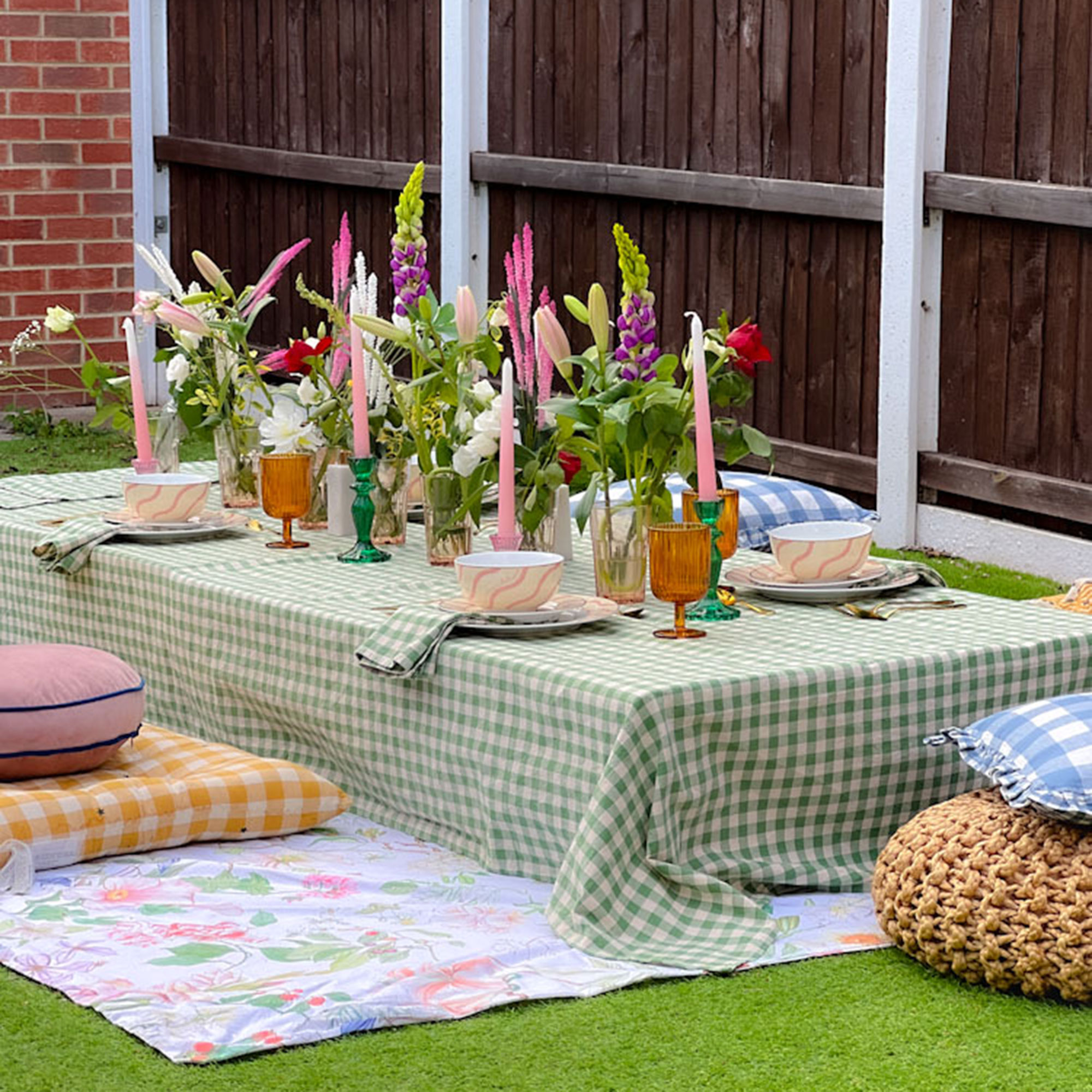 Outdoor table with green and white gingham tablecloth, flowers in tumblers alongside yellow and white gingham floor cushion and round pink pillow on top of floral ground covering