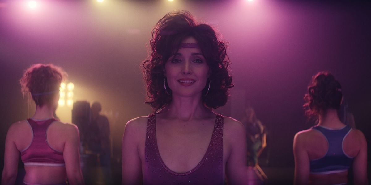 Rose Byrne with a bouncy hairdo and silver hoop earrings, wearing a sequinned purple leotard, as Sheila Rubin stands in a brightly lit aerobics studio to shoot a fitness video