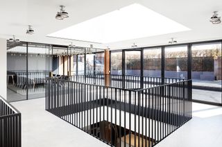 View of the upper level at Dunhill's HQ featuring light coloured flooring, multiple lights, doors that lead to the outdoor terrace and black balustrading around a rectangle opening in the floor. The boardroom can also be seen