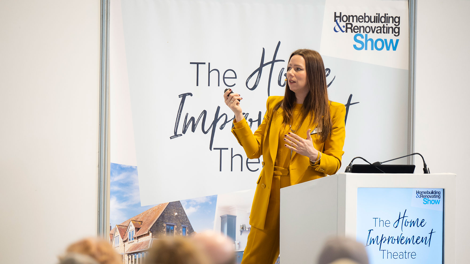 woman in a yellow suit speaking on a stage at the Homebuilding &amp; Renovating Show