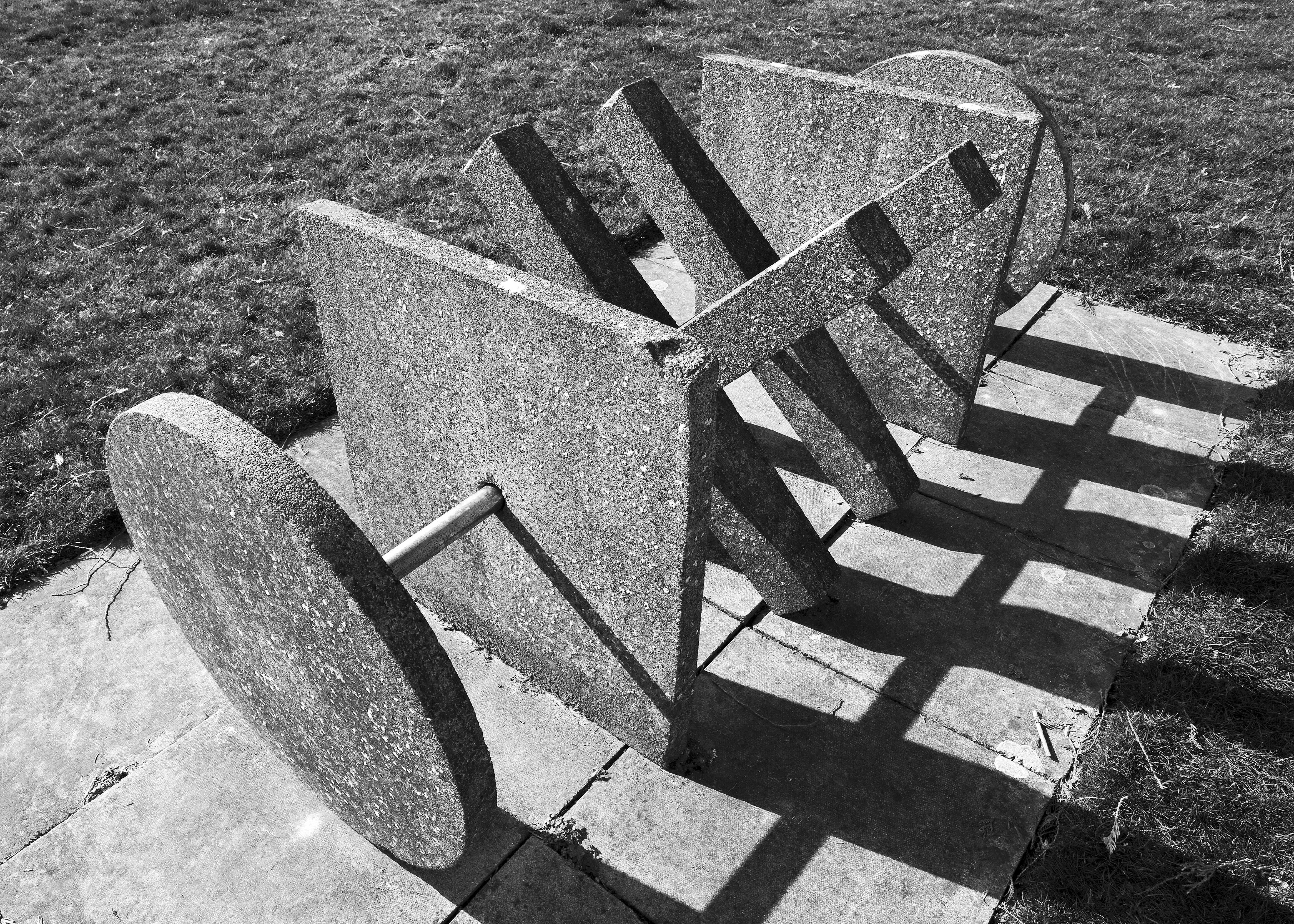 A black & white photo of a concrete sculpture. A circle, square, and x shapes are connected with a metal pole.