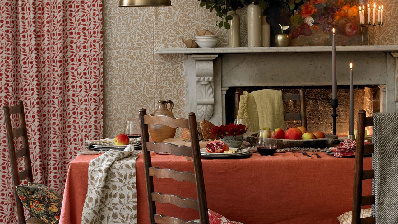 An autumnal dining room in candle light with a burnt orange tablecloth on the table and botanical beige wallpaper on the walls