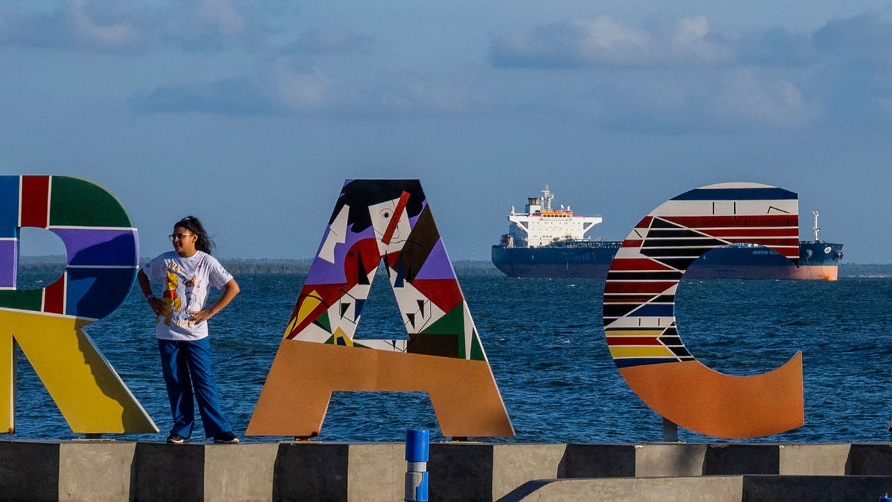 A woman poses in front of a sign welcoming visitors to Maracaibo, with an oil tanker in the background
