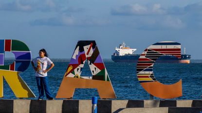 A woman poses in front of a sign welcoming visitors to Maracaibo, with an oil tanker in the background