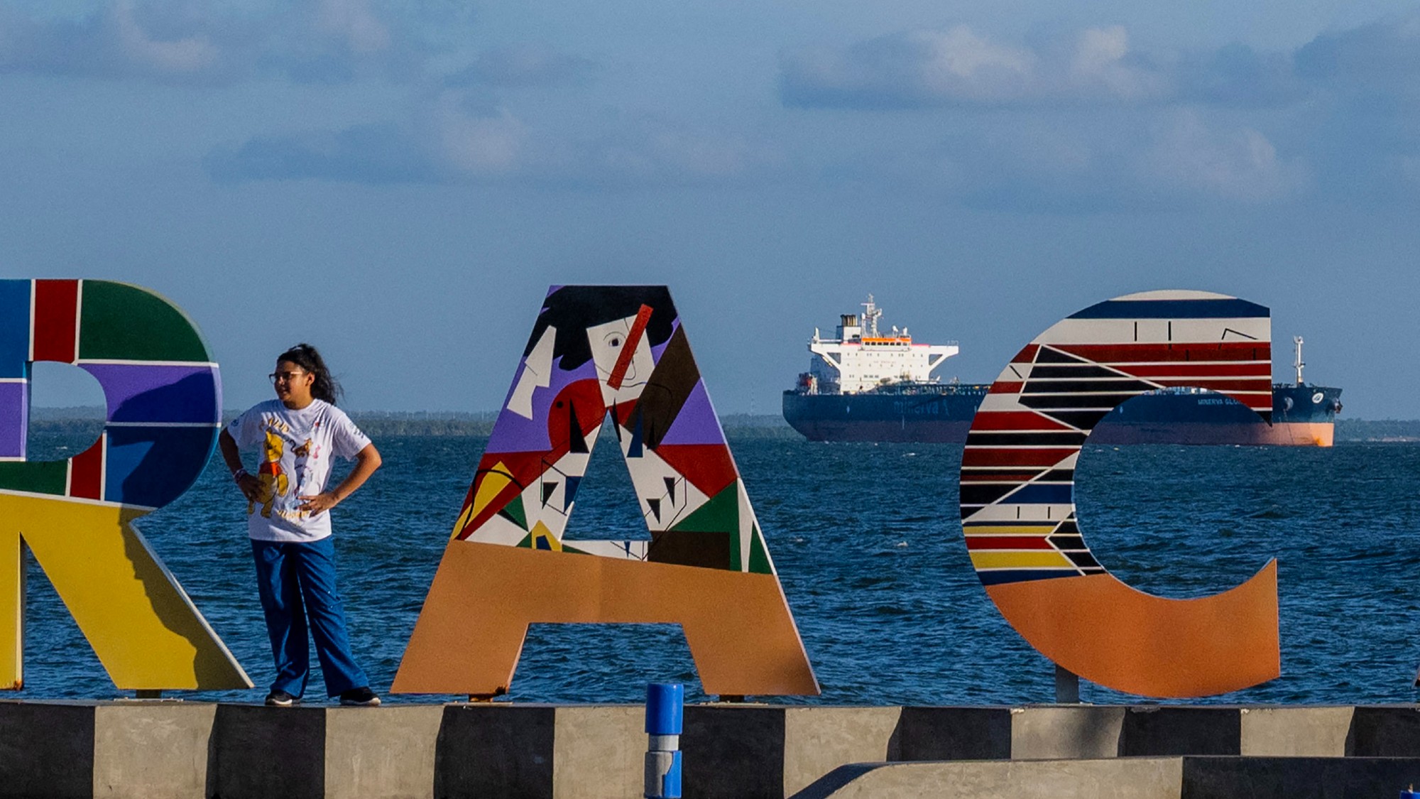 A woman poses in front of a sign welcoming visitors to Maracaibo, with an oil tanker in the background