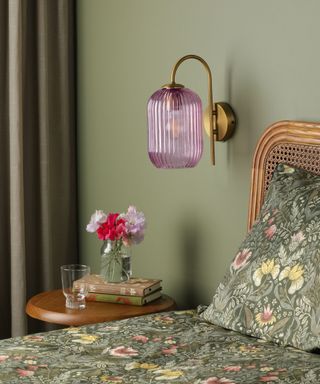 A sage green bedroom with a floral green bed and pillows, a wooden side table with books on top of it, and a purple glass light with a bronze base