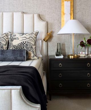 A bedroom with a white bed with two throw pillows and a black blanket on it, a black set of drawers with a white lamp and two glasses of water on top of it and a rectangular gold wall sconce above it