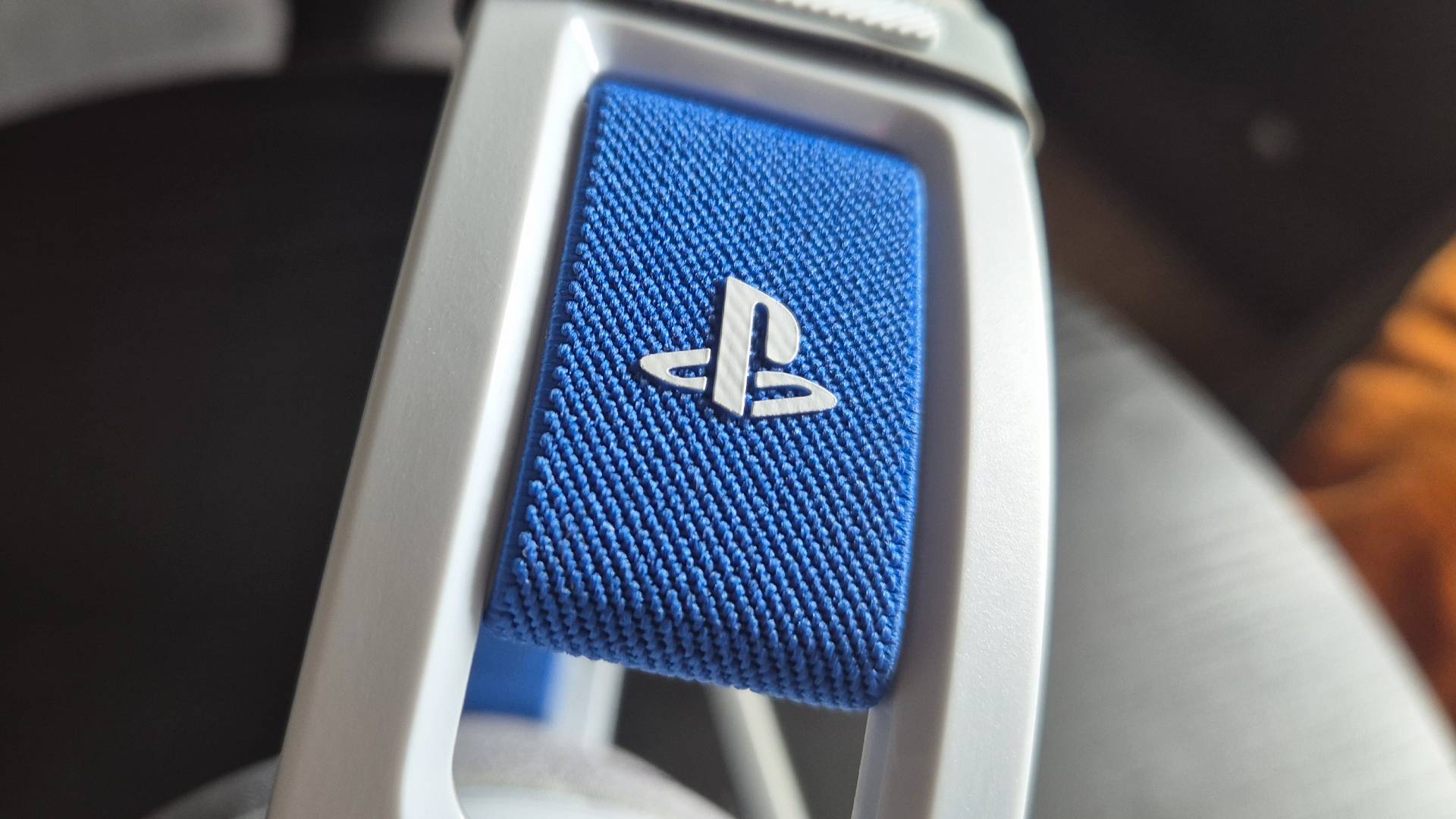 Photo of a close up of a PlayStation logo on a blue strap on a headset, with a black sidetable blurred in the background.